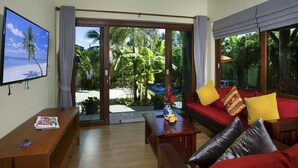 Una sala de estar con una mesa de centro de madera, sofá rojo con cojines amarillos y negros, un televisor montado en la pared y vistas a un jardín tropical a través de grandes puertas de cristal.