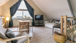A cozy loft bedroom with a sloped ceiling, a wooden desk, a TV, a comfortable sofa, and a large window with curtains. - Elegant Modern House in St Vaast la Hougue (Saint-Vaast-la-Hougue)