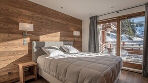 A bedroom with a wooden headboard, a bed with white bedding, a nightstand, and a view of a snowy outdoor area through large windows. - Ski-in/Ski-out Apartment with Panoramic Views and Luxury Amenities in Méribel (Méribel)