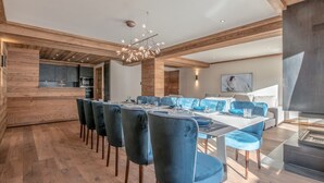 A modern dining area with a long table and blue upholstered chairs, wooden flooring, and a contemporary chandelier. - Ski-in/Ski-out Apartment with Panoramic Views and Luxury Amenities in Méribel (Méribel)