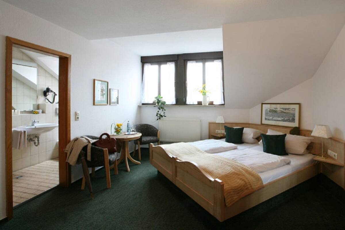 Een hotelkamer met een bed, een stoel, een klein tafeltje, een raam met gordijnen, een radiator en een spiegel.
