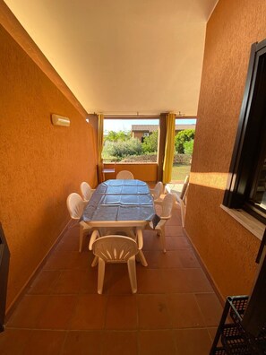 Eine überdachte Terrasse mit Tisch und Stühlen, ein Fenster mit Vorhängen und Blick auf grüne Vegetation.