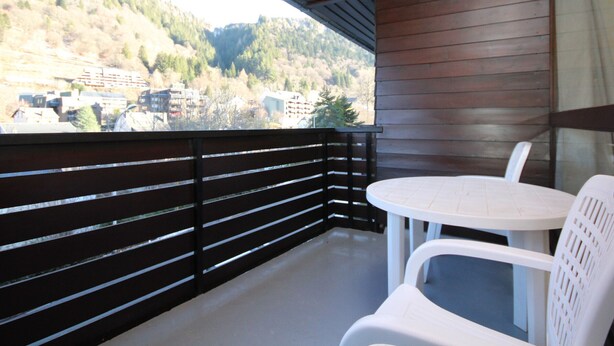 A balcony with a wooden railing, a round table, and two white plastic chairs. - Mont-Dore: 4BR Duplex with Balcony, Garage, and Wi-Fi (Mont-Dore)