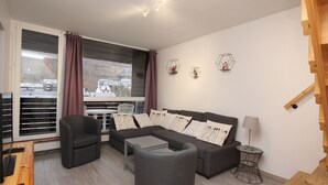 A modern living room with a grey sofa, a black armchair, and a small table. A window with blinds and curtains is visible, and there are decorative wall pieces above the sofa. - Mont-Dore: 4BR Duplex with Balcony, Garage, and Wi-Fi (Mont-Dore)