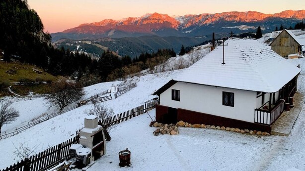 A snow-covered house with a wooden fence, a chimney, and a view of mountains in the background. - Charming Traditional House in a Pristine Location (Fundata)