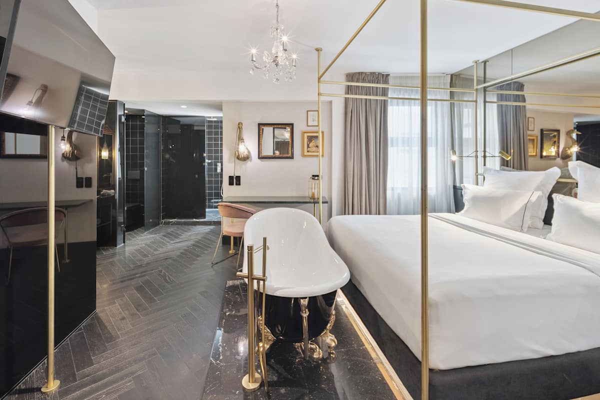 A modern hotel room with a large bed, a sitting area with a chair and a small table, a mirror, and a chandelier.