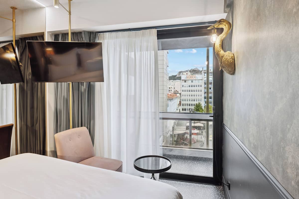 A modern hotel room with a bed, a round table, a chair, and a large window offering a city view.