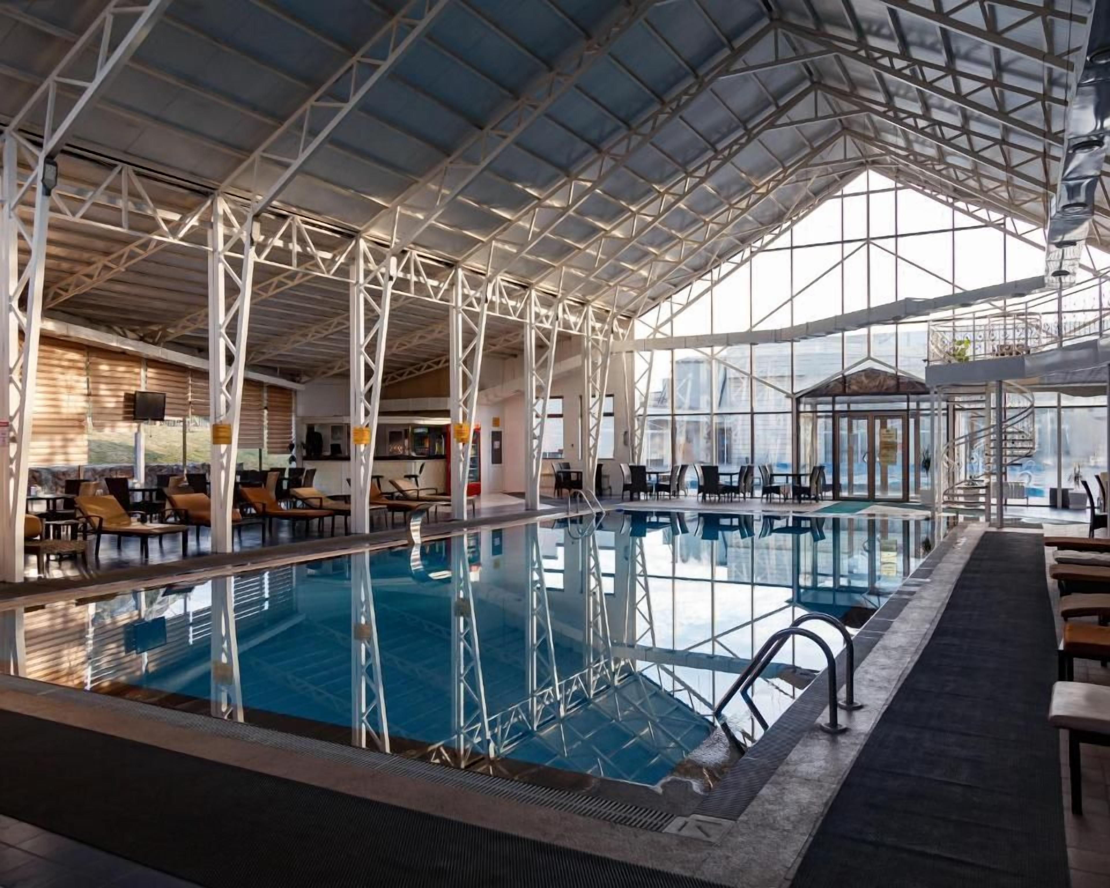 Een binnenzwembad met een hoge plafond en glazen wanden, omringd door ligbedden en tafels.