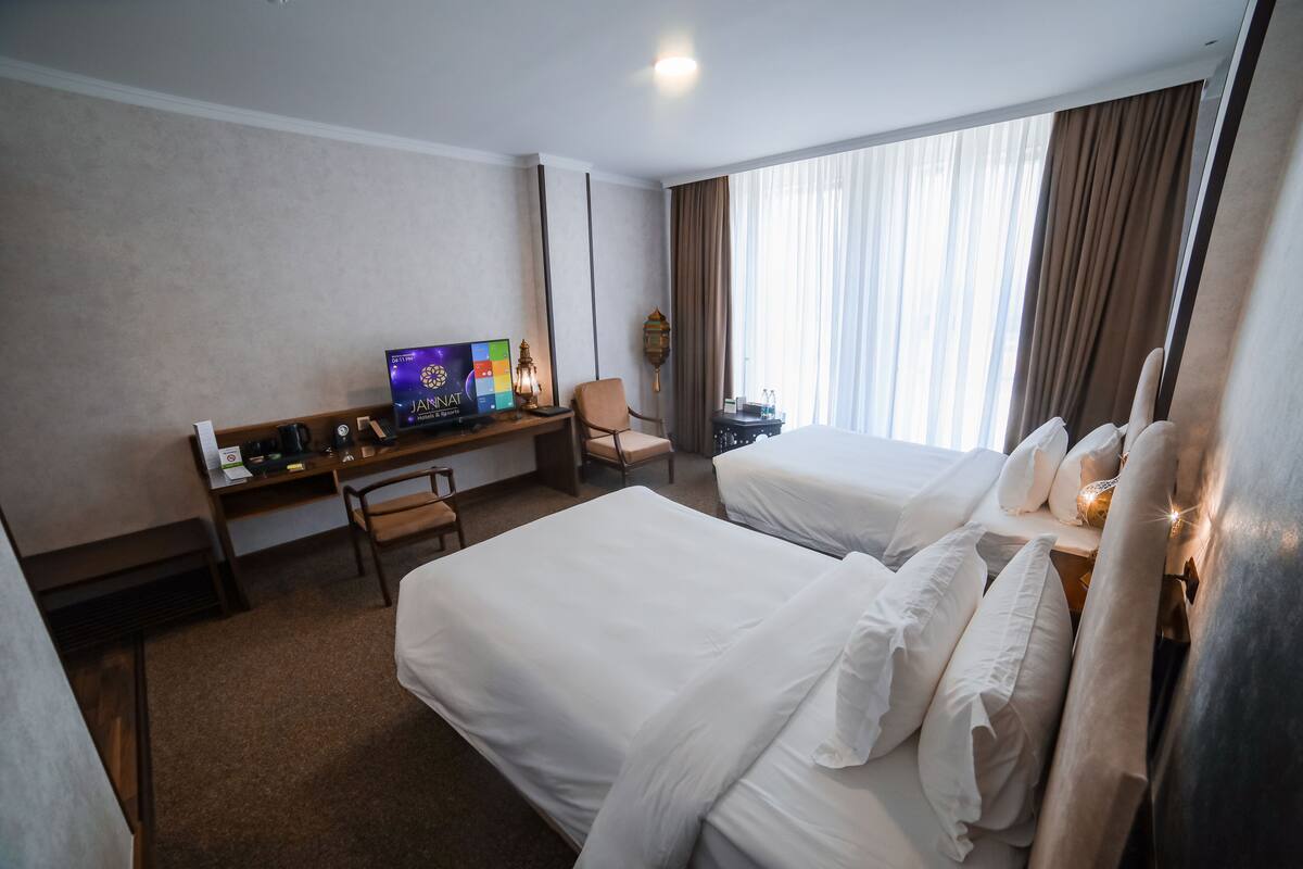 A hotel room with two beds, a desk with a computer, a chair, and a large window with curtains.