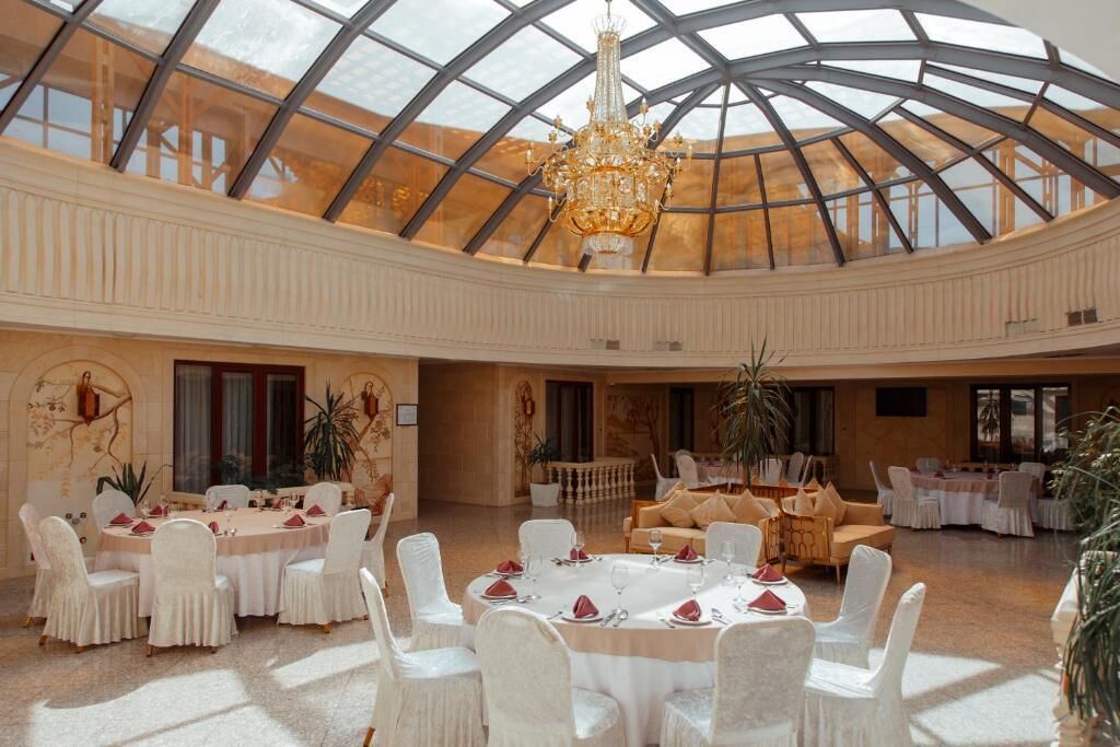 A spacious hall with a high ceiling and a glass dome, featuring round tables with white chairs and a chandelier.