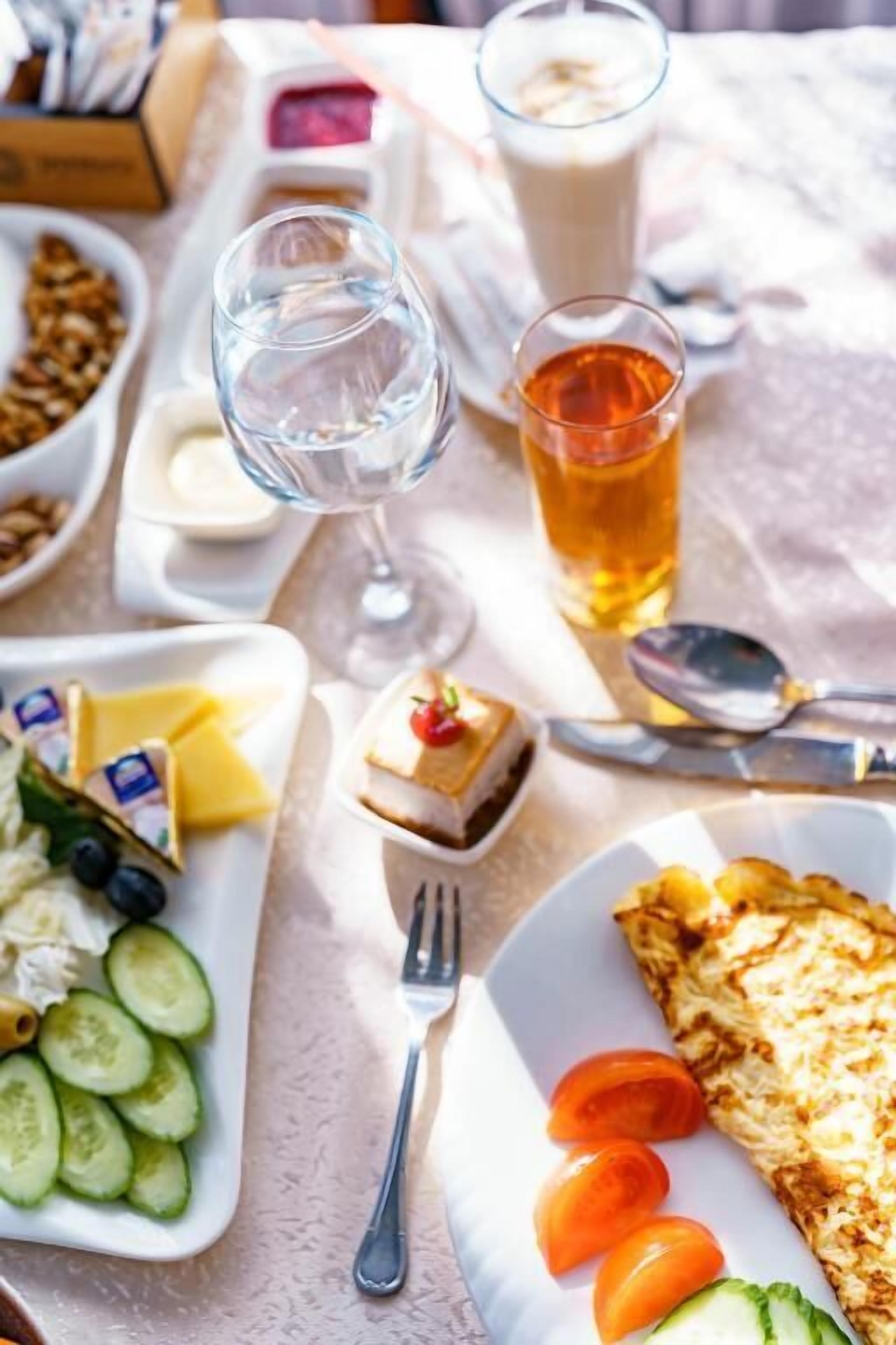 Une table est dressée avec différents aliments, notamment une assiette de tomates en tranches, des concombres, du fromage et un petit dessert. On y trouve également deux verres, l’un contenant un liquide transparent et l’autre un liquide doré, qui pourrait être du thé.