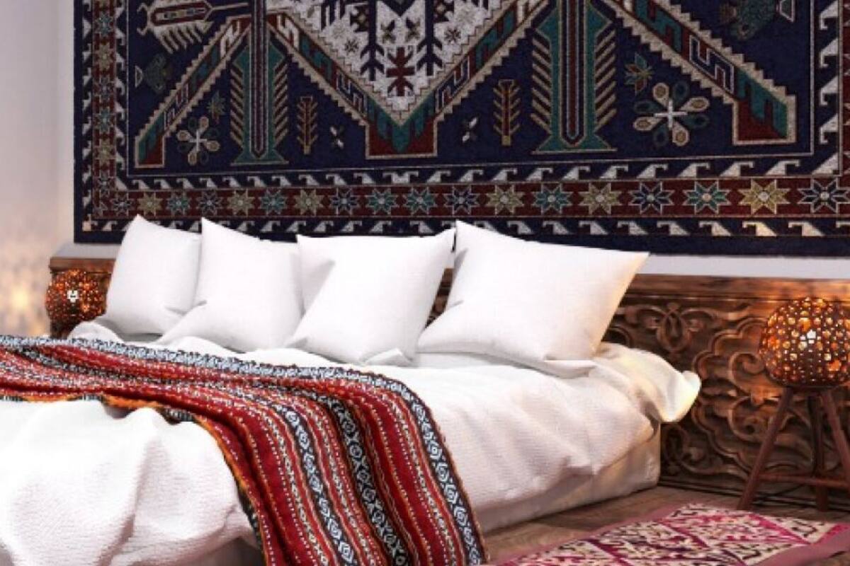 A bed with white bedding and a red patterned blanket, a wooden nightstand with a cup and a book, and a decorative tapestry on the wall.