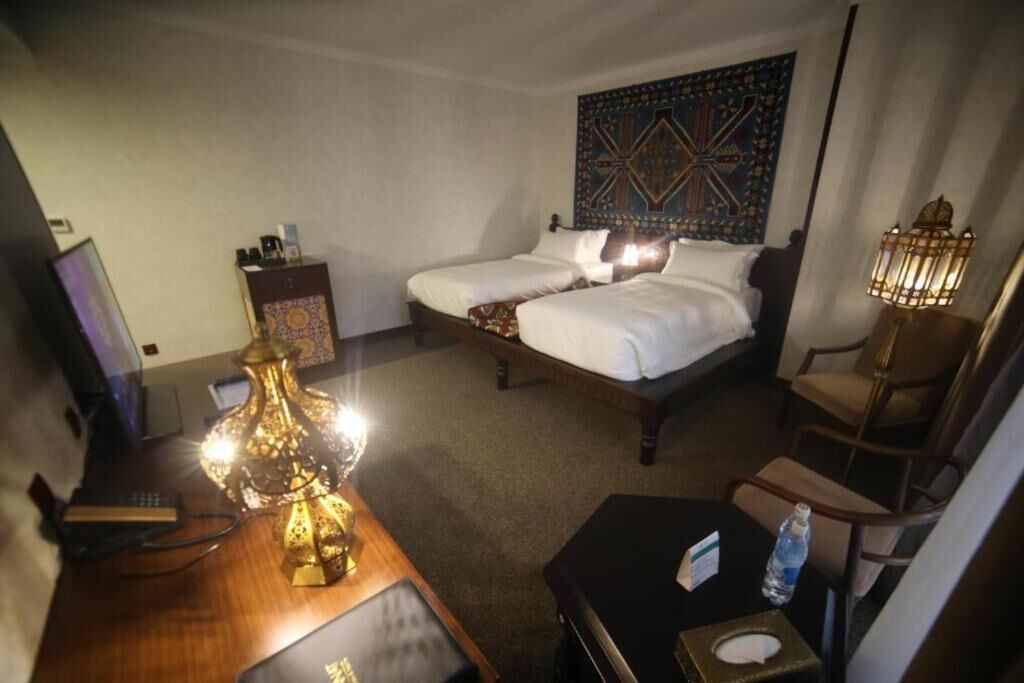 A hotel room with two beds, a desk with a lamp, a chair, a small table with a bottle, and a wall tapestry.