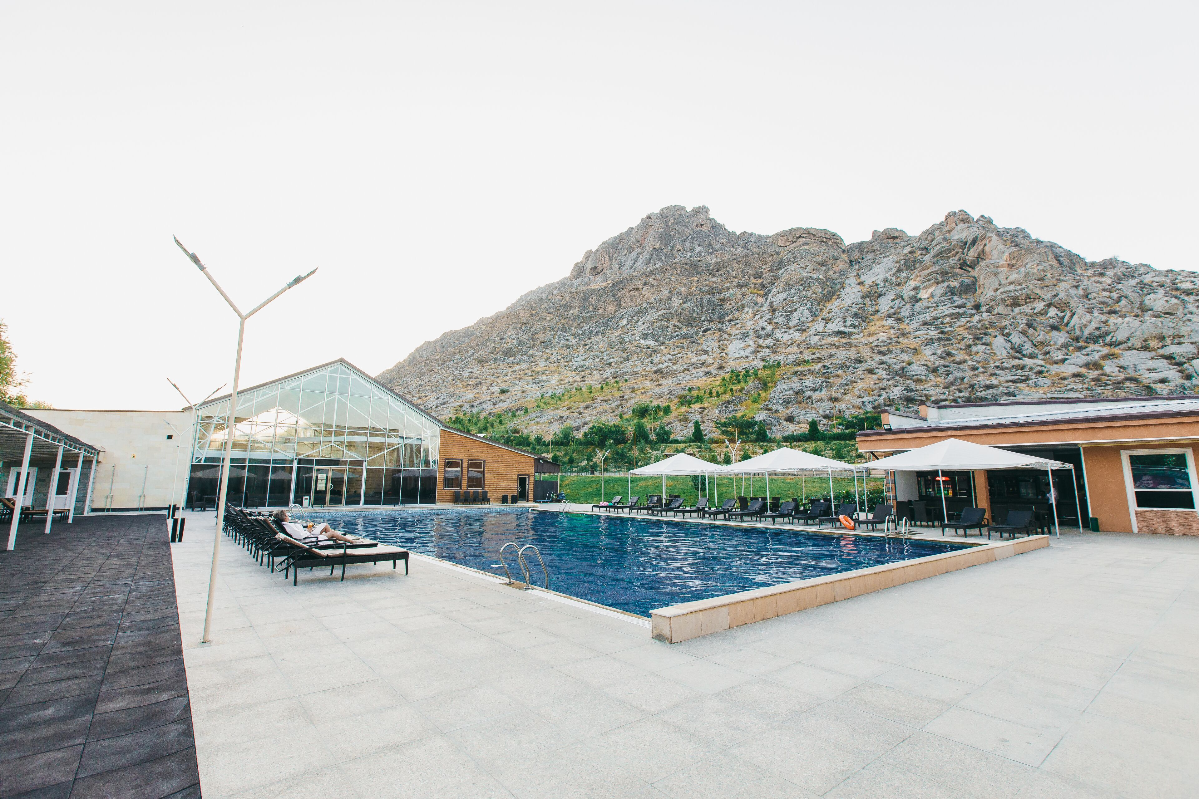 Una piscina con tumbonas y sombrillas, un edificio de cristal y montañas al fondo.