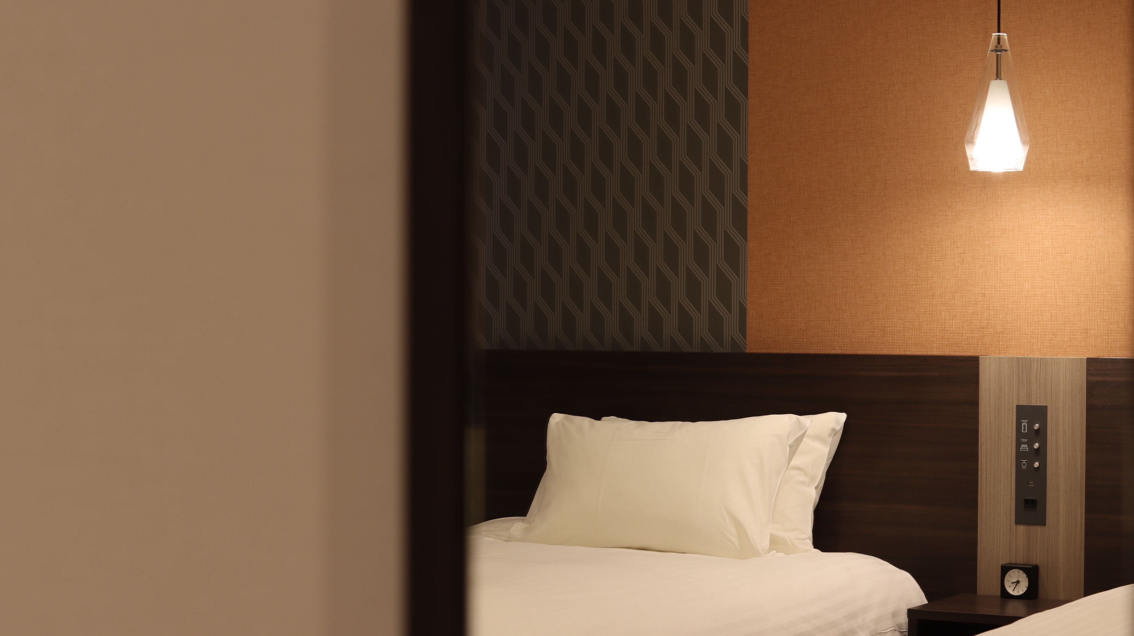 A hotel room with a bed, a nightstand, a wall clock, and a control panel.