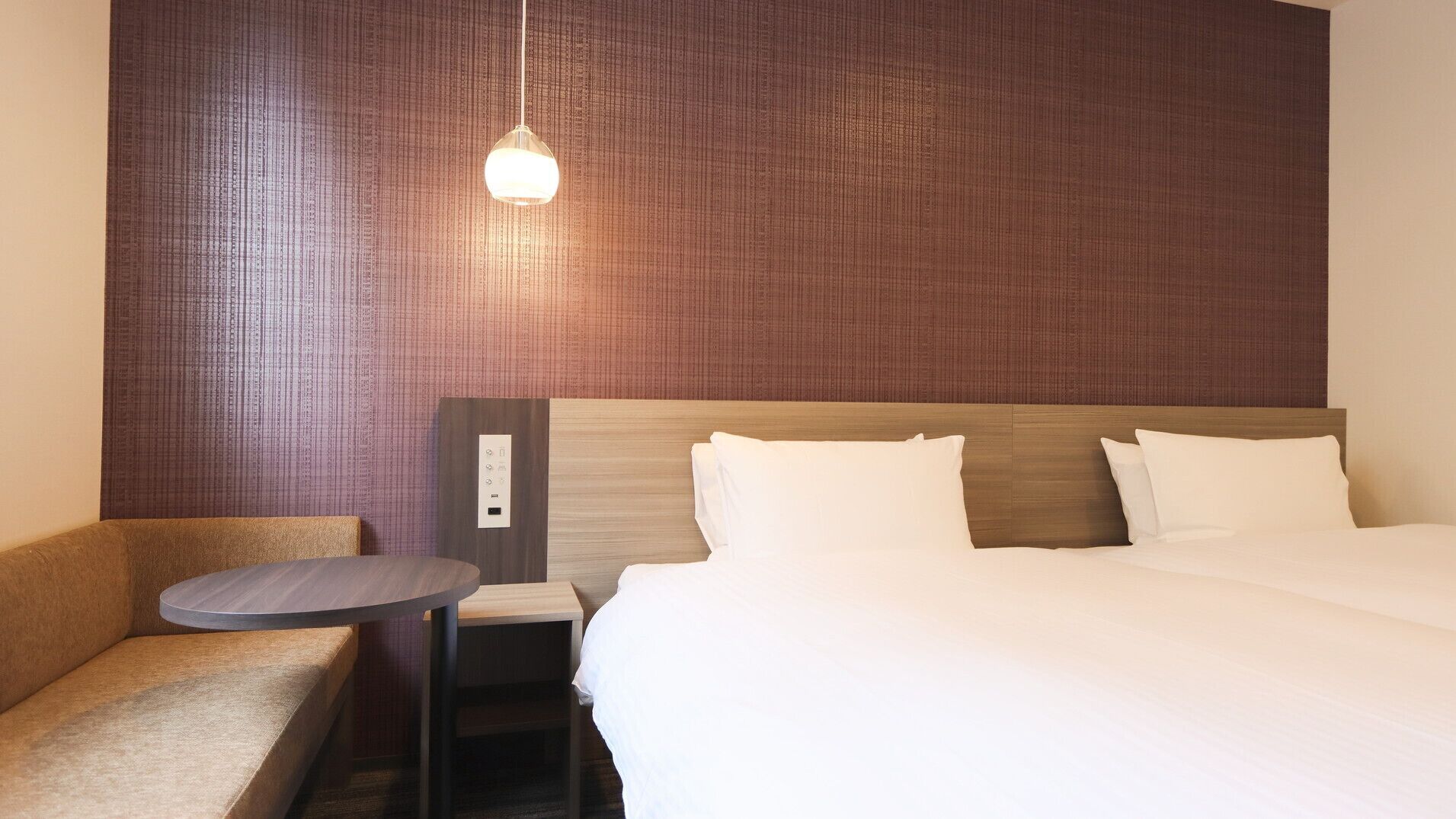 Une chambre d’hôtel comprenant un lit, une table de chevet, une chaise et un panneau de commande fixé au mur.