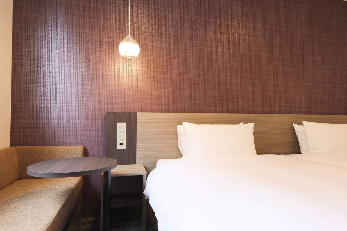 Une chambre d’hôtel comprenant un lit, une table de chevet, une chaise et un panneau de commande fixé au mur.