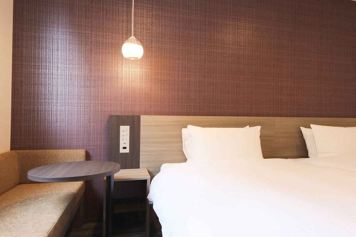 A hotel room with a bed, a bedside table, a chair, and a wall-mounted control panel.