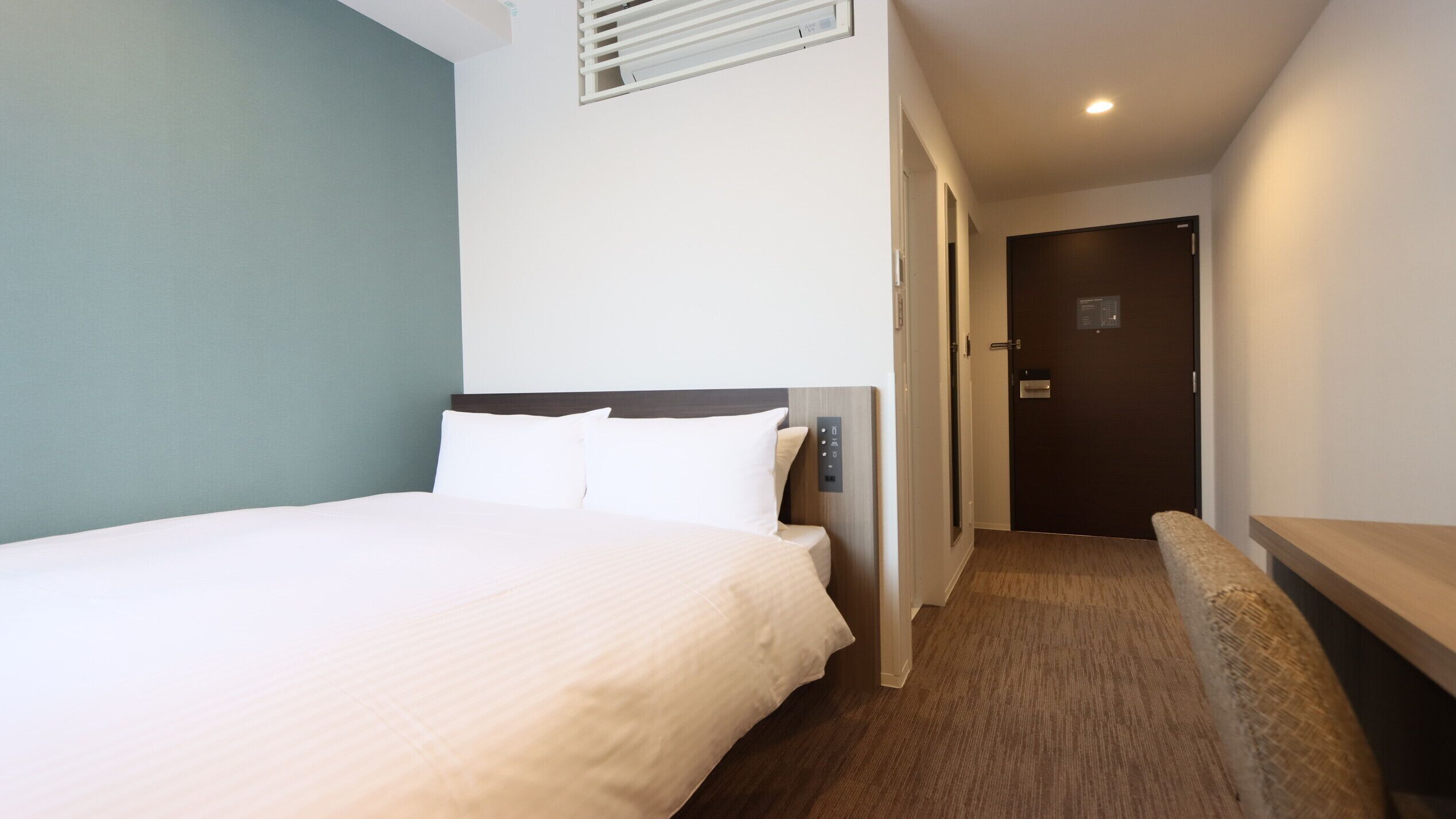 Une chambre d’hôtel comprenant un lit, un panneau de commande situé près du lit, un banc, une porte et une bouche d’aération fixée au mur.