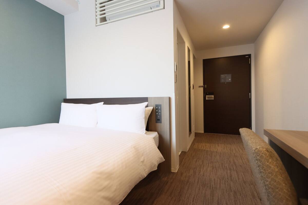 Une chambre d’hôtel comprenant un lit, un panneau de commande situé près du lit, un banc, une porte et une bouche d’aération fixée au mur.