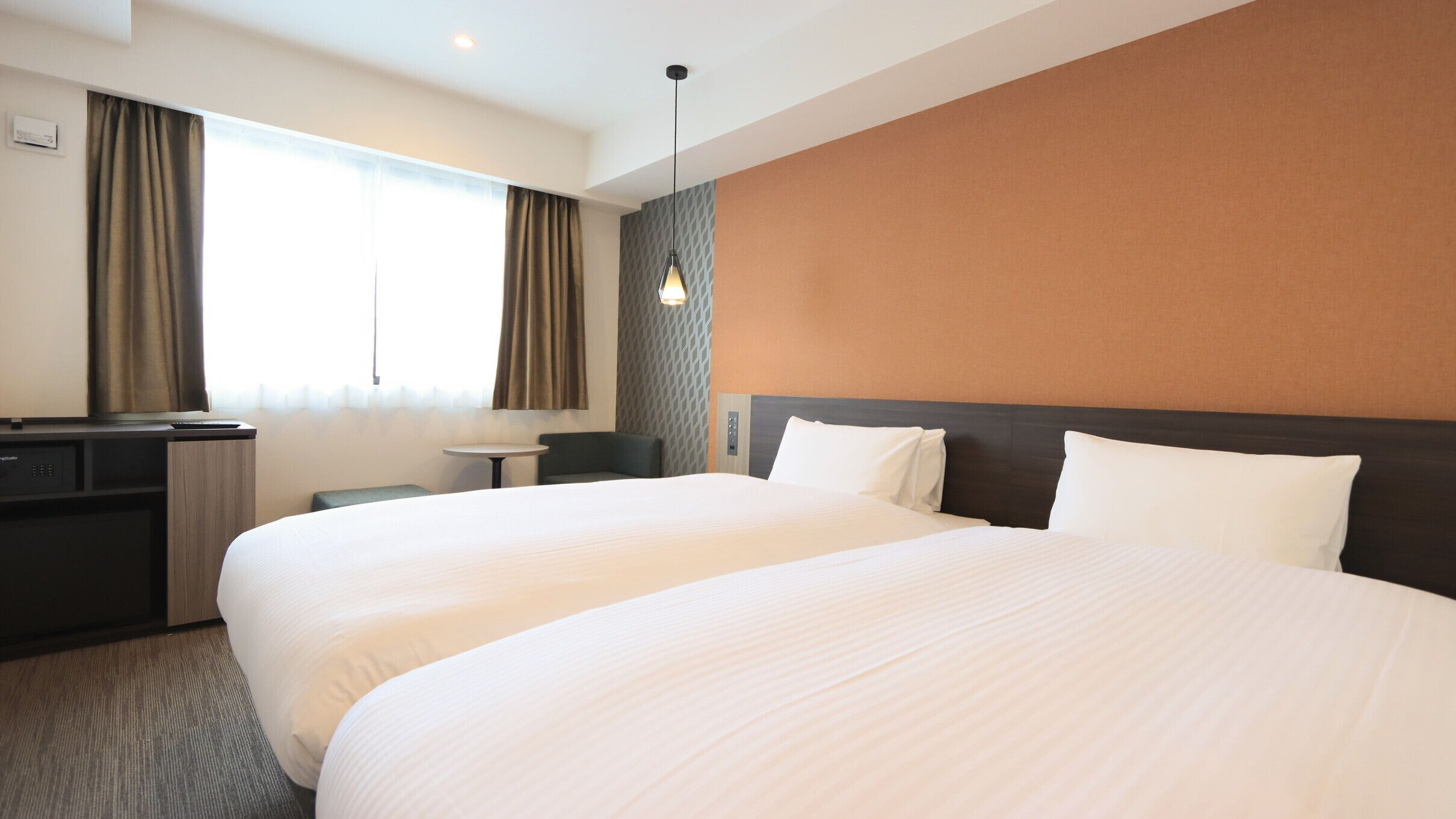 Une chambre d’hôtel avec deux lits, un bureau, une chaise, une petite table et une lampe fixée au mur.
