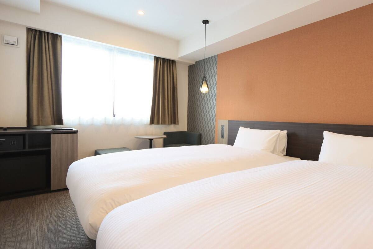 Une chambre d’hôtel avec deux lits, un bureau, une chaise, une petite table et une lampe fixée au mur.