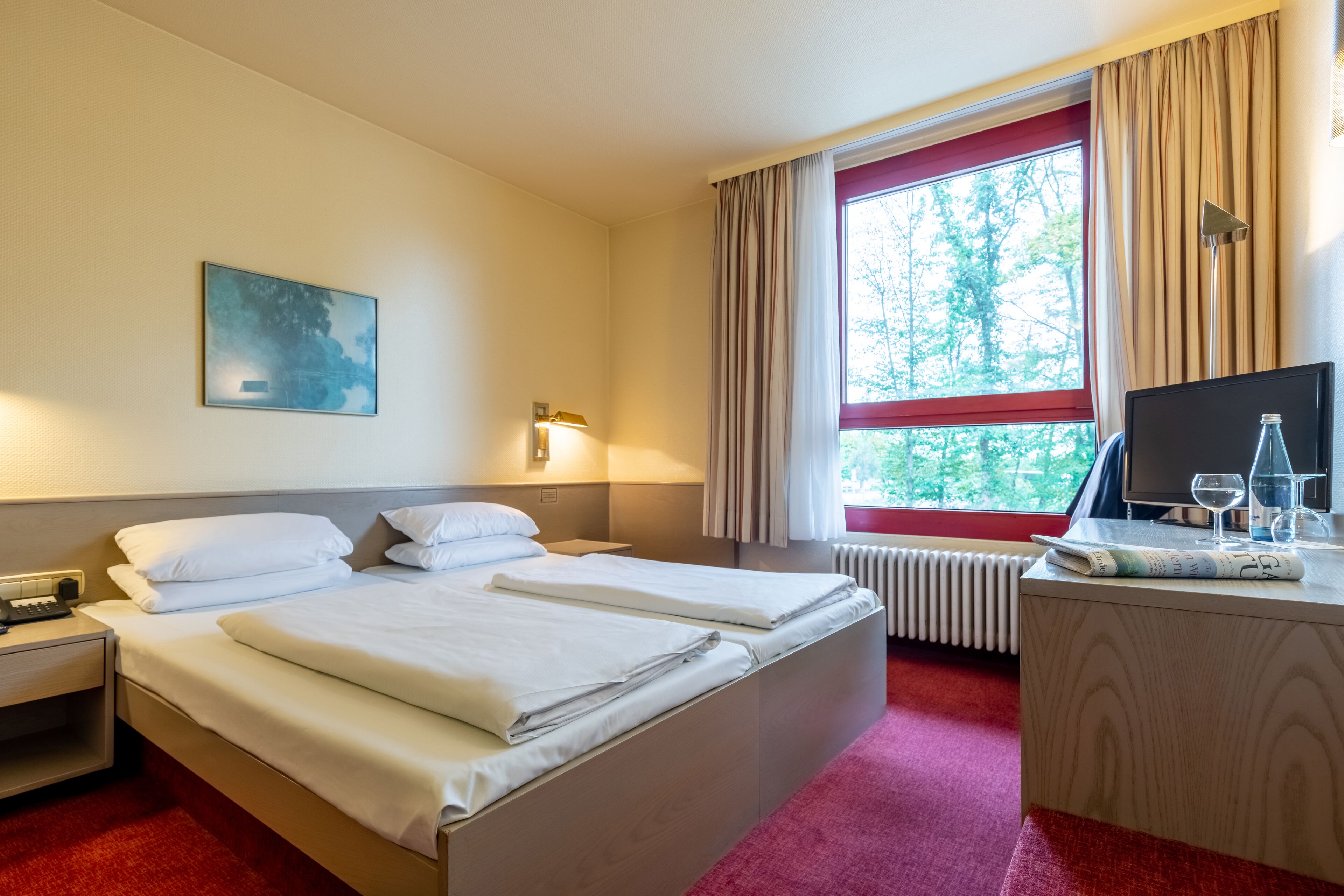 Ein Hotelzimmer mit einem Bett, einem Schreibtisch mit Computer und Brille, einem Fenster mit Vorhängen und einem Bild an der Wand.