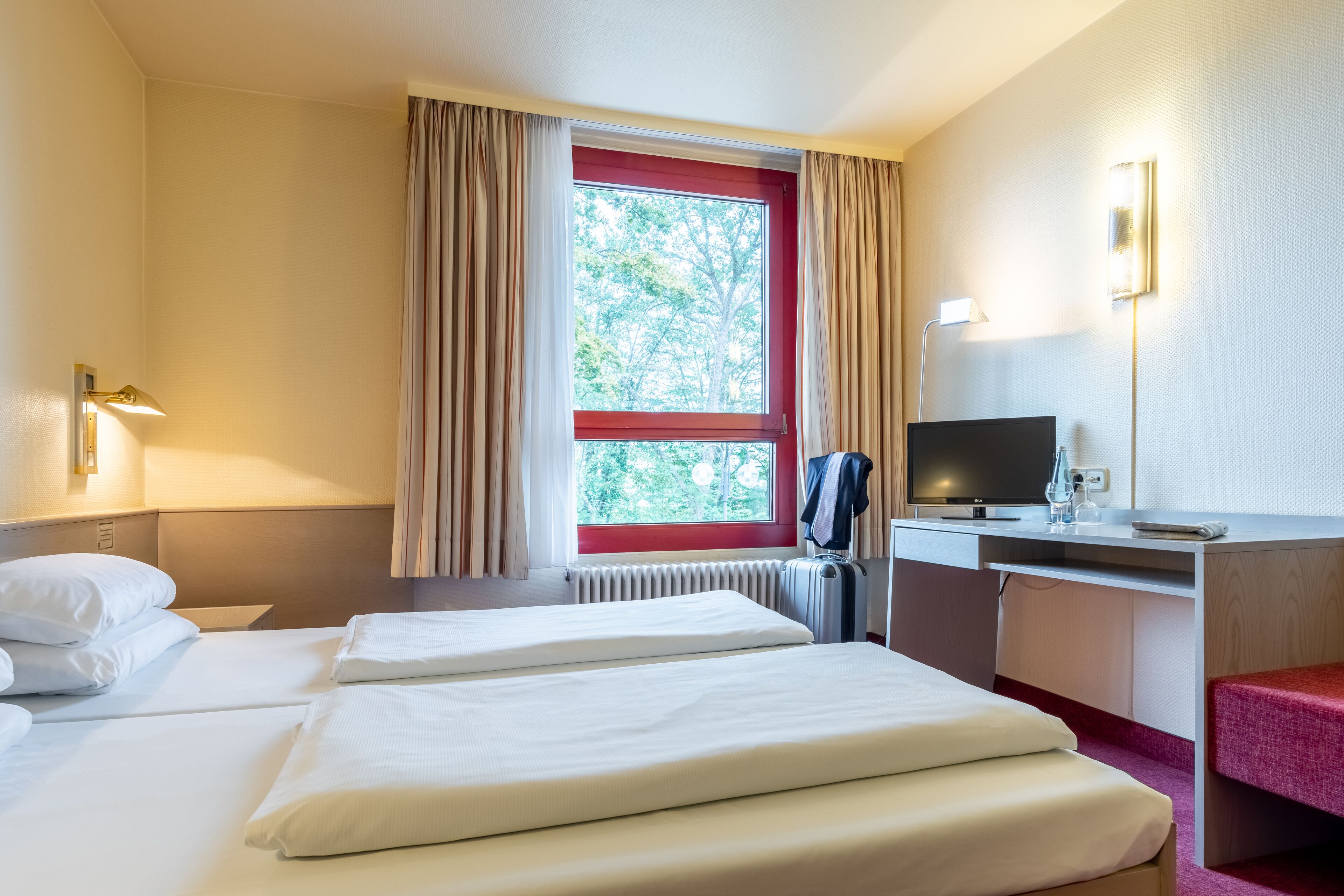A hotel room with two beds, a desk with a computer, a TV, and a window with curtains.