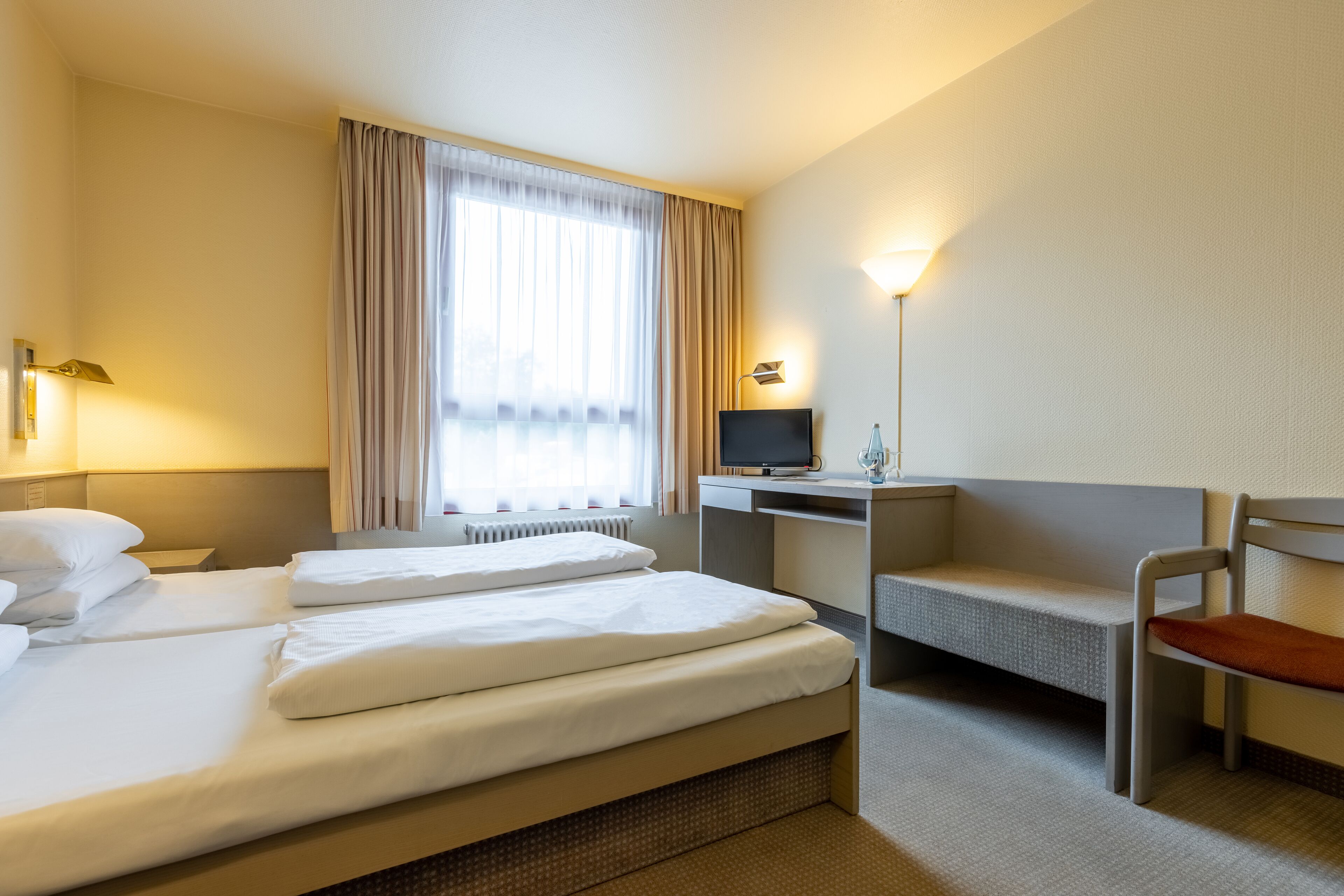 Habitación de hotel con dos camas, un escritorio, una silla, un televisor y una ventana con cortinas.