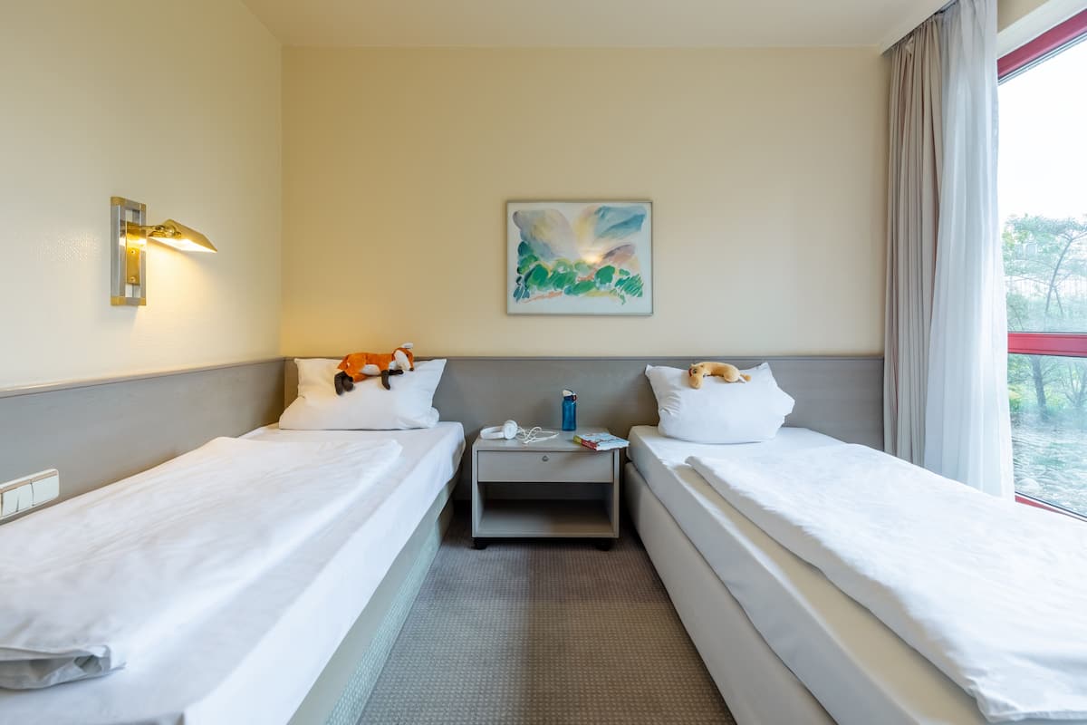 A hotel room with two single beds, a nightstand, a lamp, a painting, and a window with curtains.