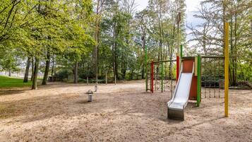 Ein Spielplatz mit Rutsche und Schaukeln, umgeben von Bäumen und einem Erdboden.