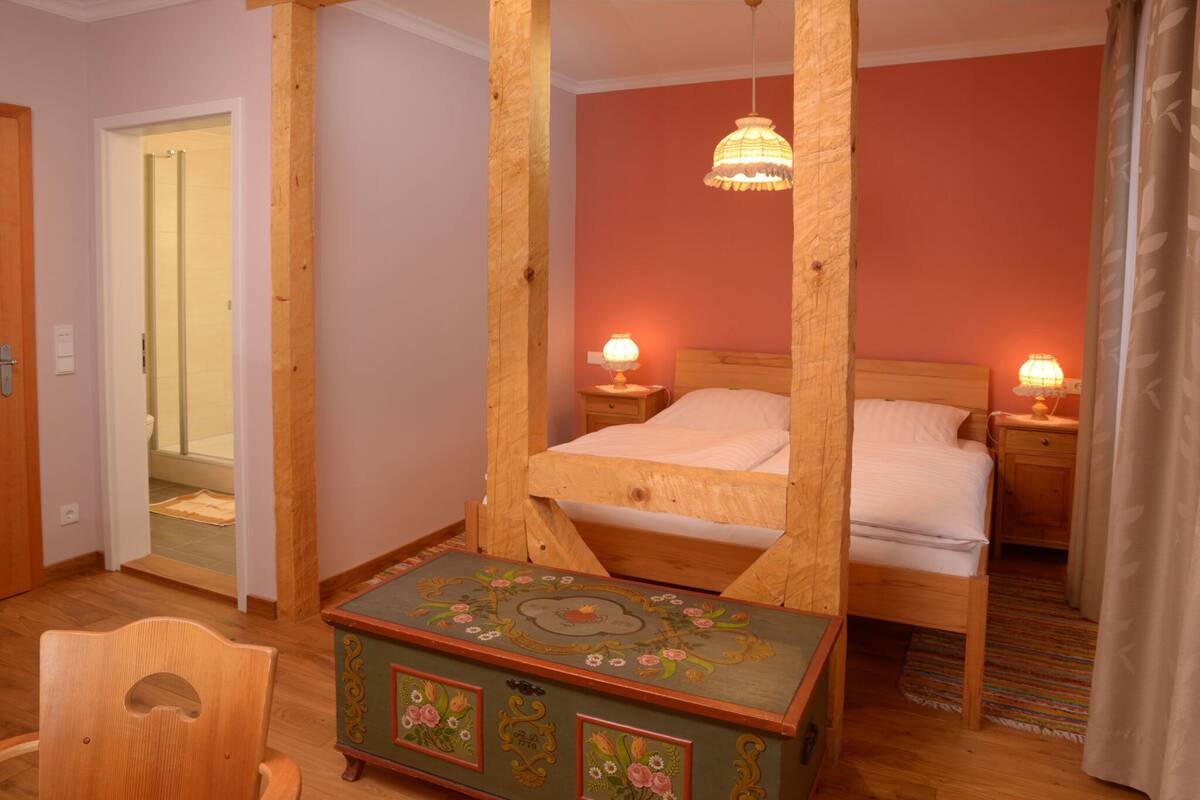 A bedroom with a wooden four-poster bed, two bedside tables with lamps, a wooden chest of drawers, and a door leading to another room.
