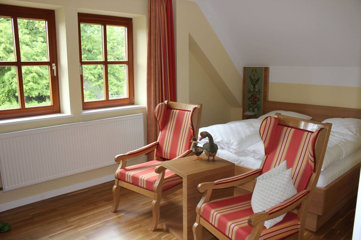 A bedroom with a bed, two striped chairs, a wooden table, and a window with curtains.