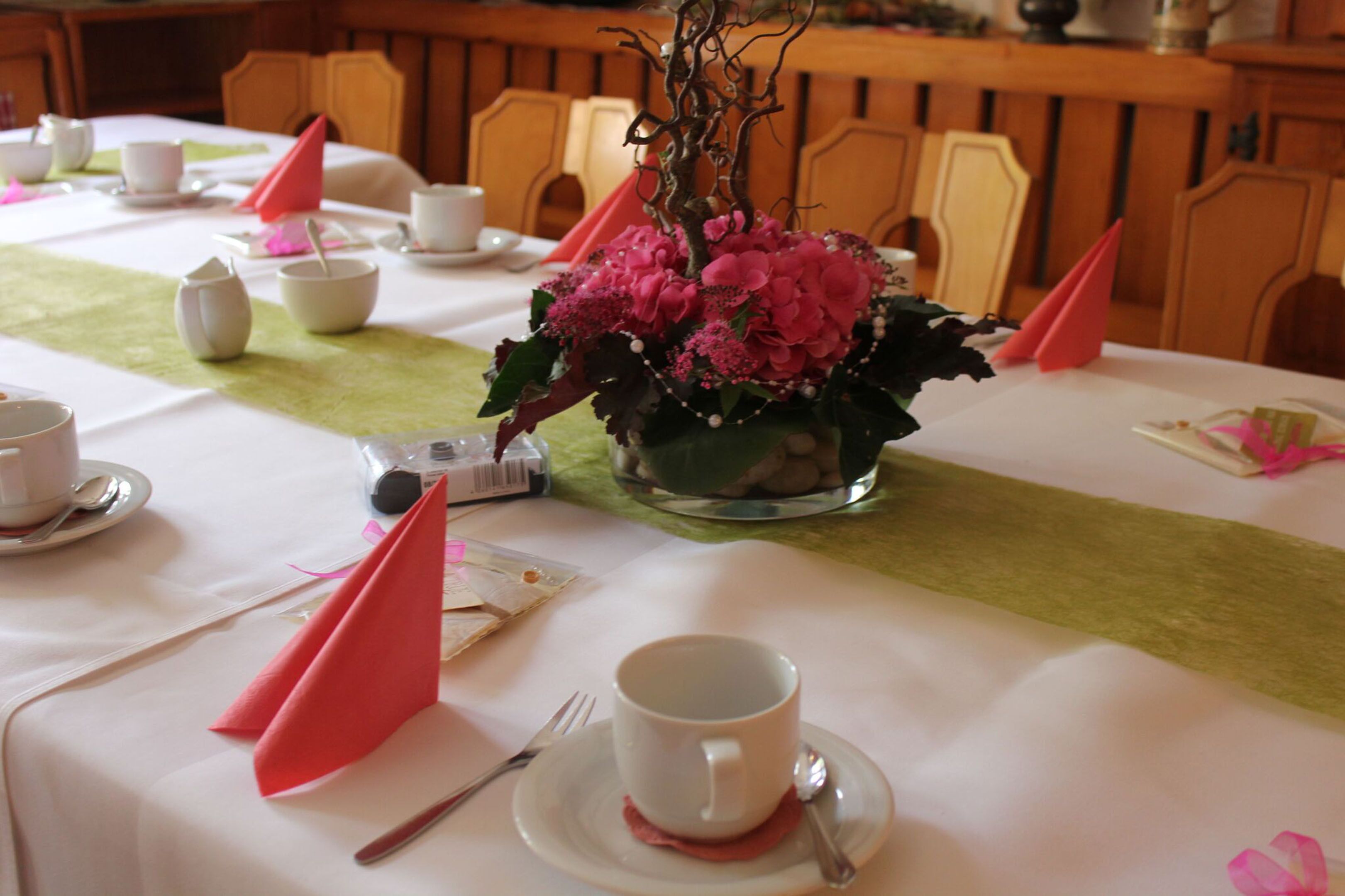 A dining table set with white tableware, a centerpiece of pink flowers, and a green table runner.