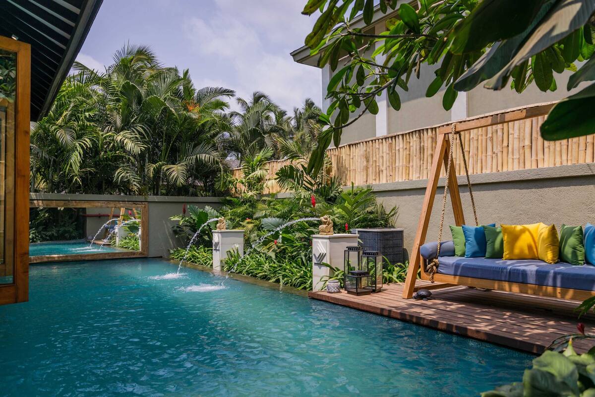 Un área de piscina con terraza de madera, una silla colgante y un sofá con cojines de colores.