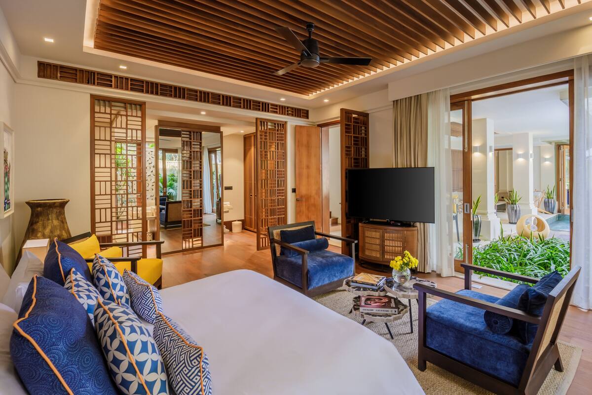 Amplia habitación de hotel con una cama grande, dos sillones, un televisor y un balcón con plantas.