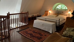 Ein Schlafzimmer mit schrägen Wänden, einem Bett, einem Nachttisch, einem Sessel, einem Fenster und einer Treppe.