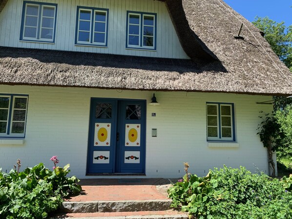 Uma casa tradicional com telhado de palha, uma porta azul com painéis decorativos e um caminho de tijolos que leva à entrada.