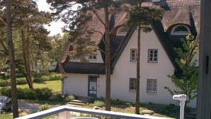 Ein Haus mit weißer Fassade, dunklem Dach und blauer Tür. Im Vordergrund ein weißer Zaun und ein Stuhl mit gestreifter Sitzauflage.