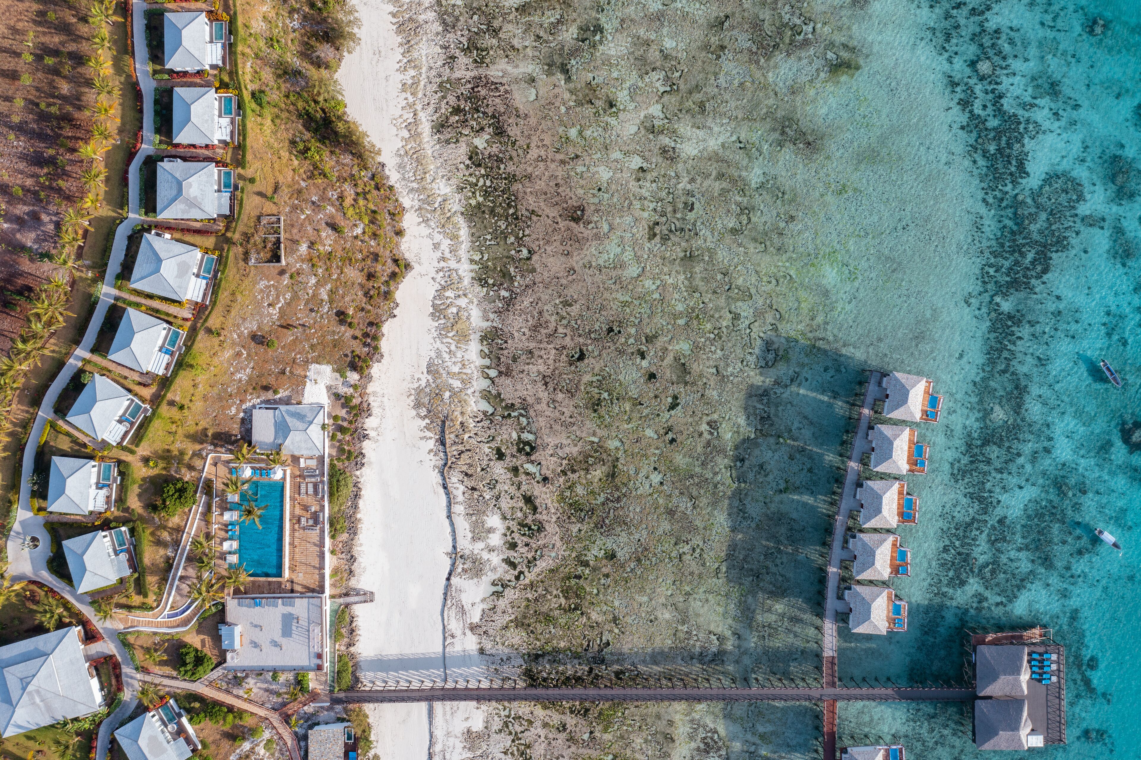 Vista aérea de un complejo turístico costero con varias cabañas sobre el agua, una piscina central y un puente que conecta las edificaciones con tierra firme.