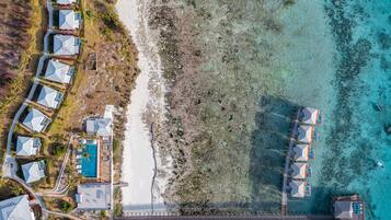 Vista aérea de un complejo turístico costero con varias cabañas sobre el agua, una piscina central y un puente que conecta las edificaciones con tierra firme.