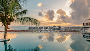 Un área de piscina tranquila con sillones de descanso, una palmera y bungalows sobre el agua al fondo.