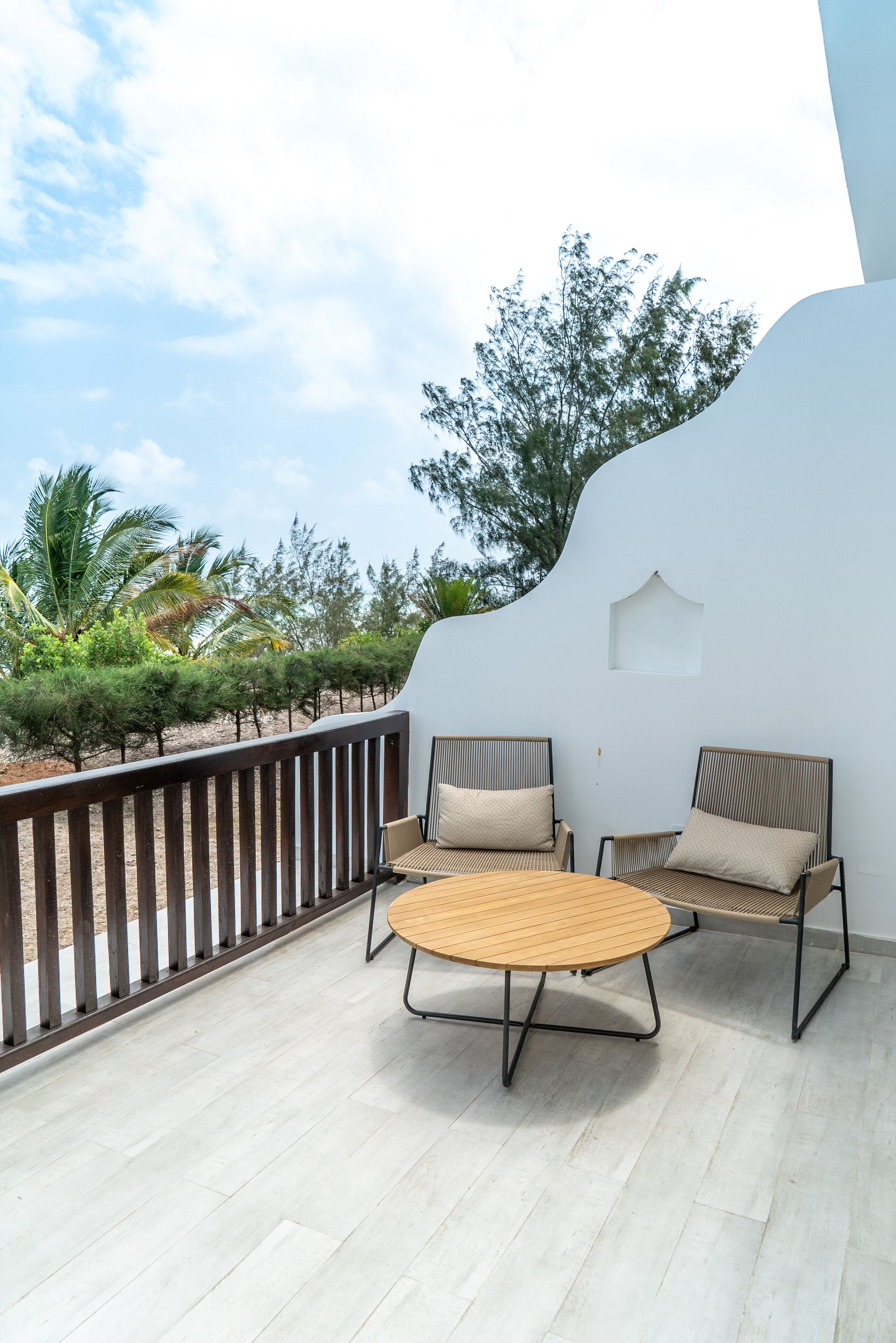 A balcony with a round wooden table and two chairs, surrounded by a white wall and a wooden railing.