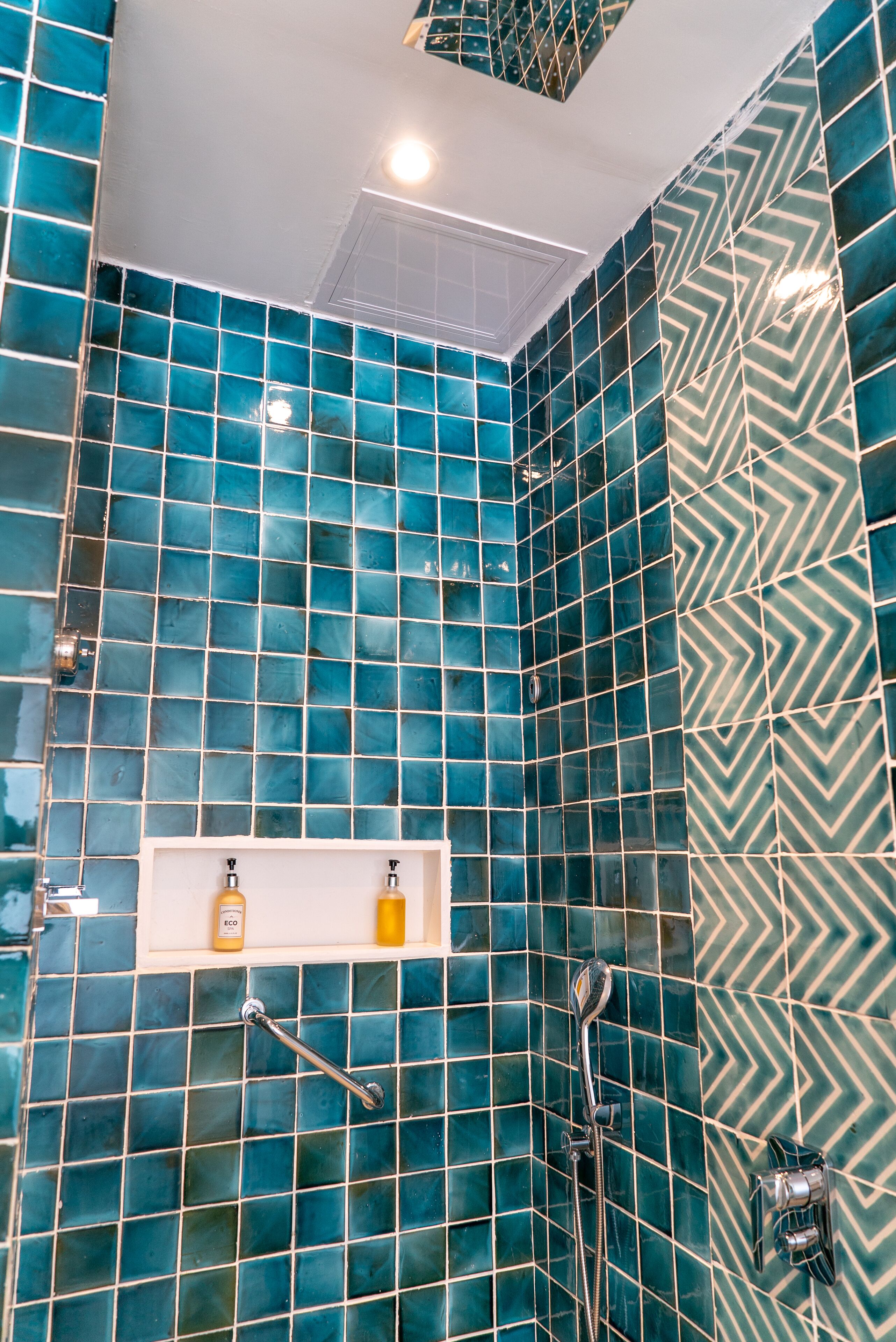 Un baño con mosaicos azules, un estante blanco con artículos de aseo y una ducha con cabezal de ducha de mano.