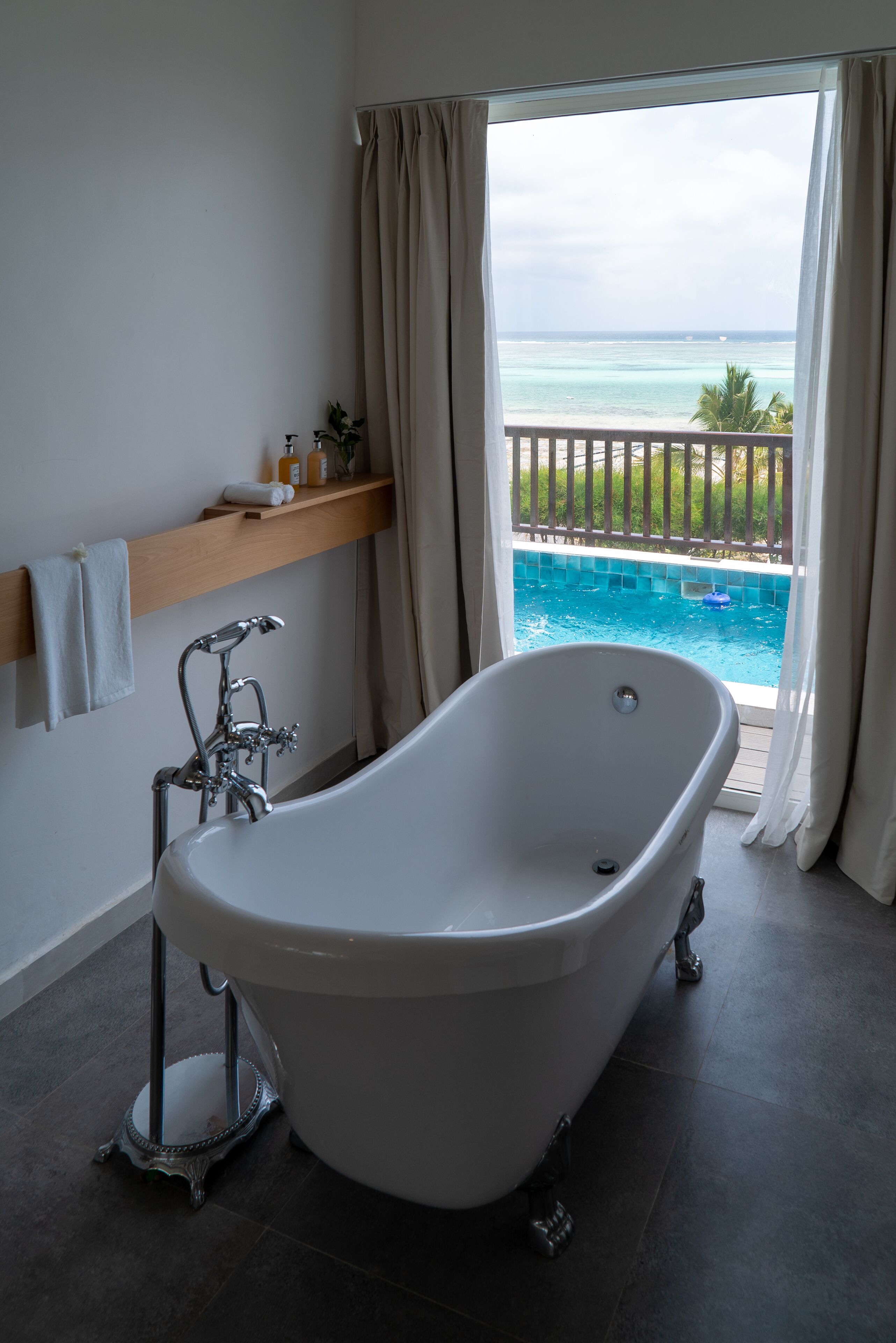 A bathroom with a freestanding bathtub, a wooden shelf with toiletries, and a view of a pool and ocean through a large window.