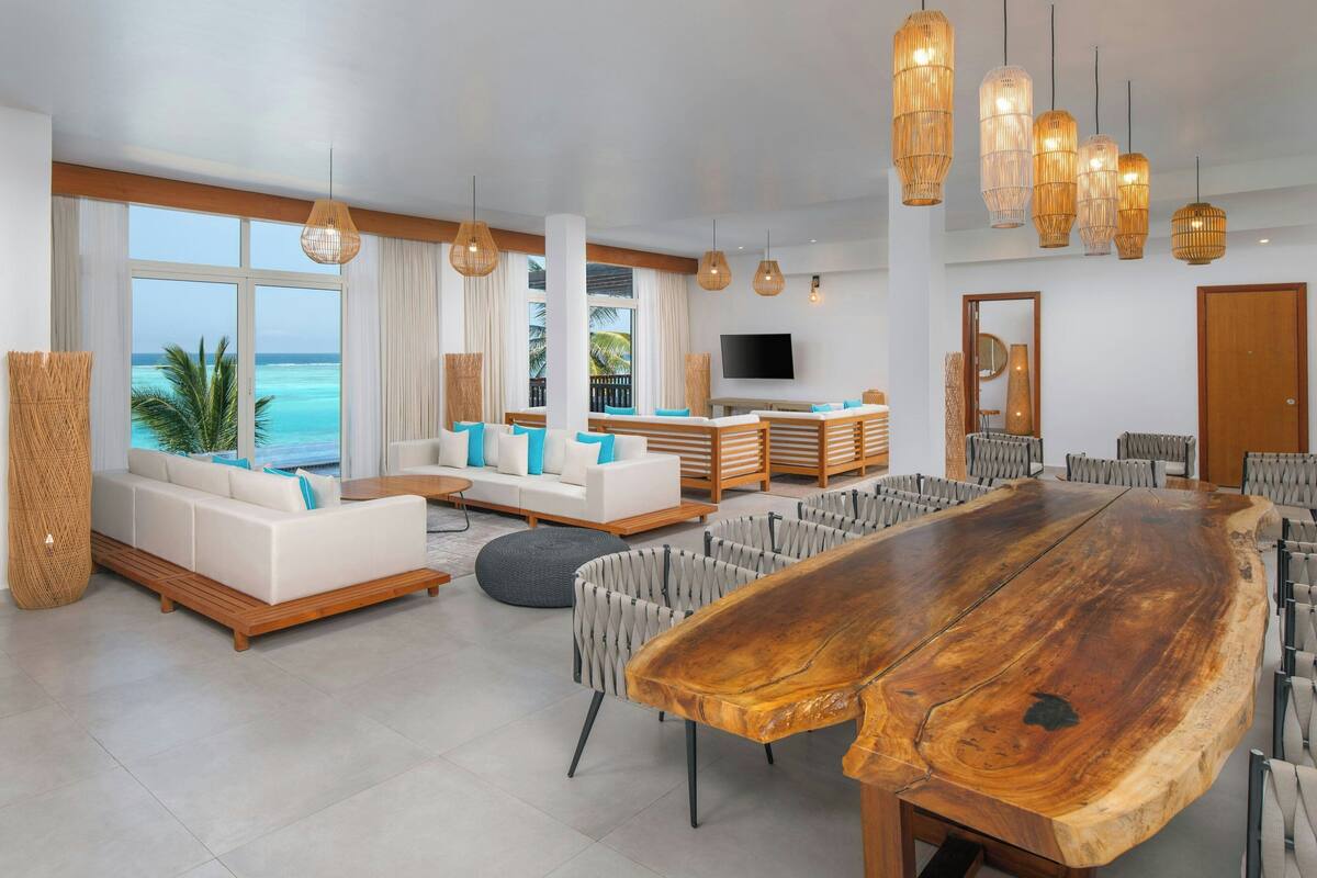 A spacious living area with a large wooden dining table, white sofas with blue cushions, and a view of the ocean.