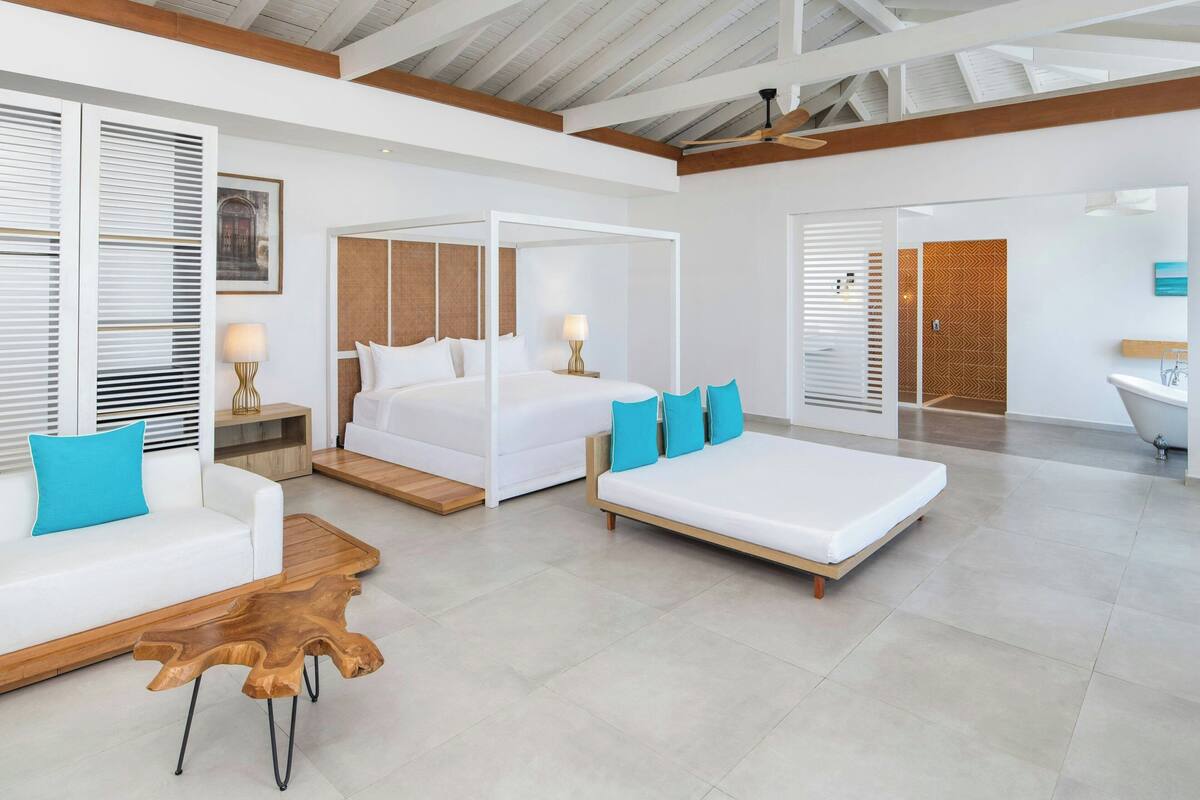 A modern hotel room with a four-poster bed, a white sofa, a wooden coffee table, and a bathroom with a bathtub and sink.
