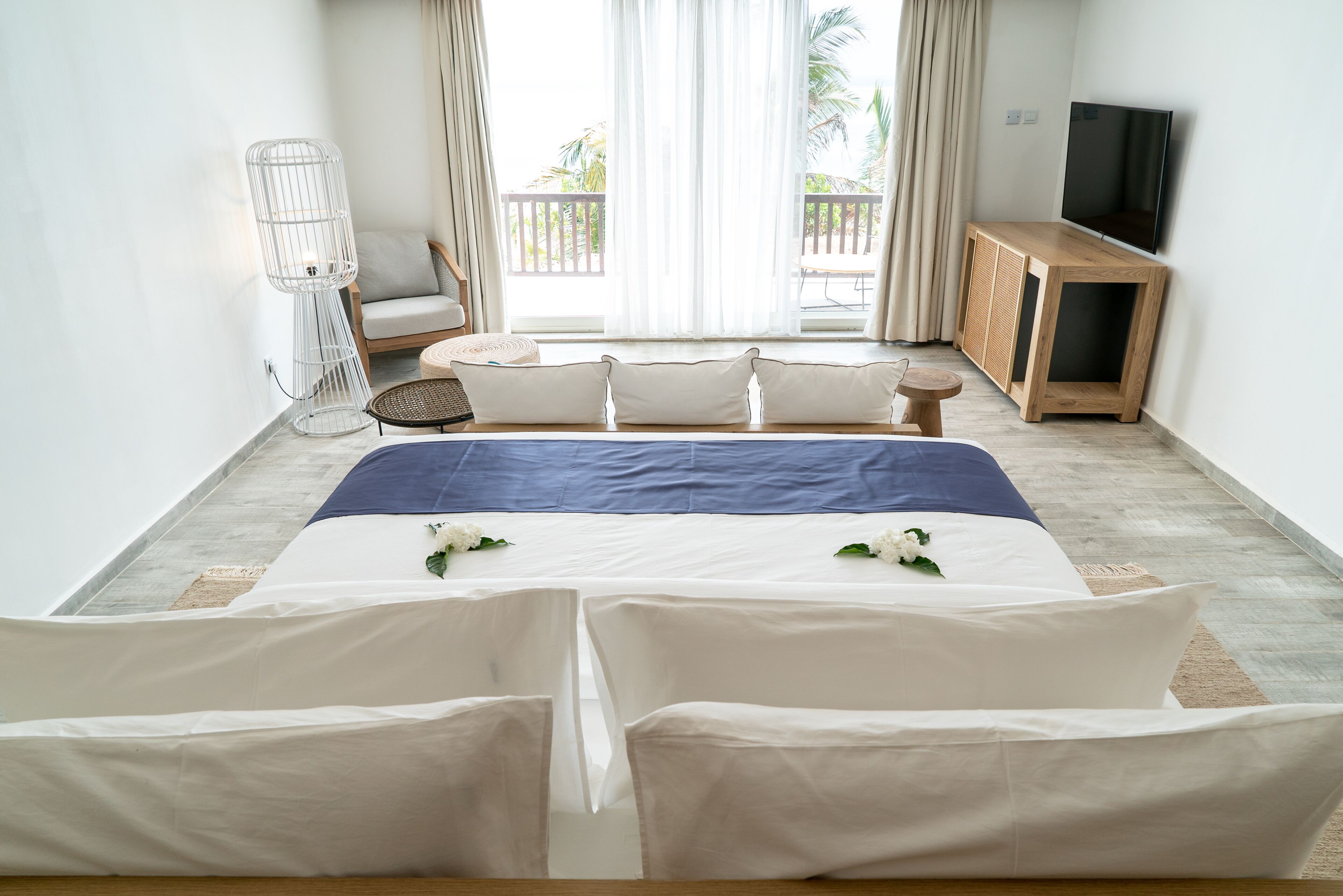 Une chambre d’hôtel moderne dotée d’un grand lit, d’une télévision, d’un coin salon avec un fauteuil et une petite table, ainsi que d’un balcon offrant une vue sur les palmiers.