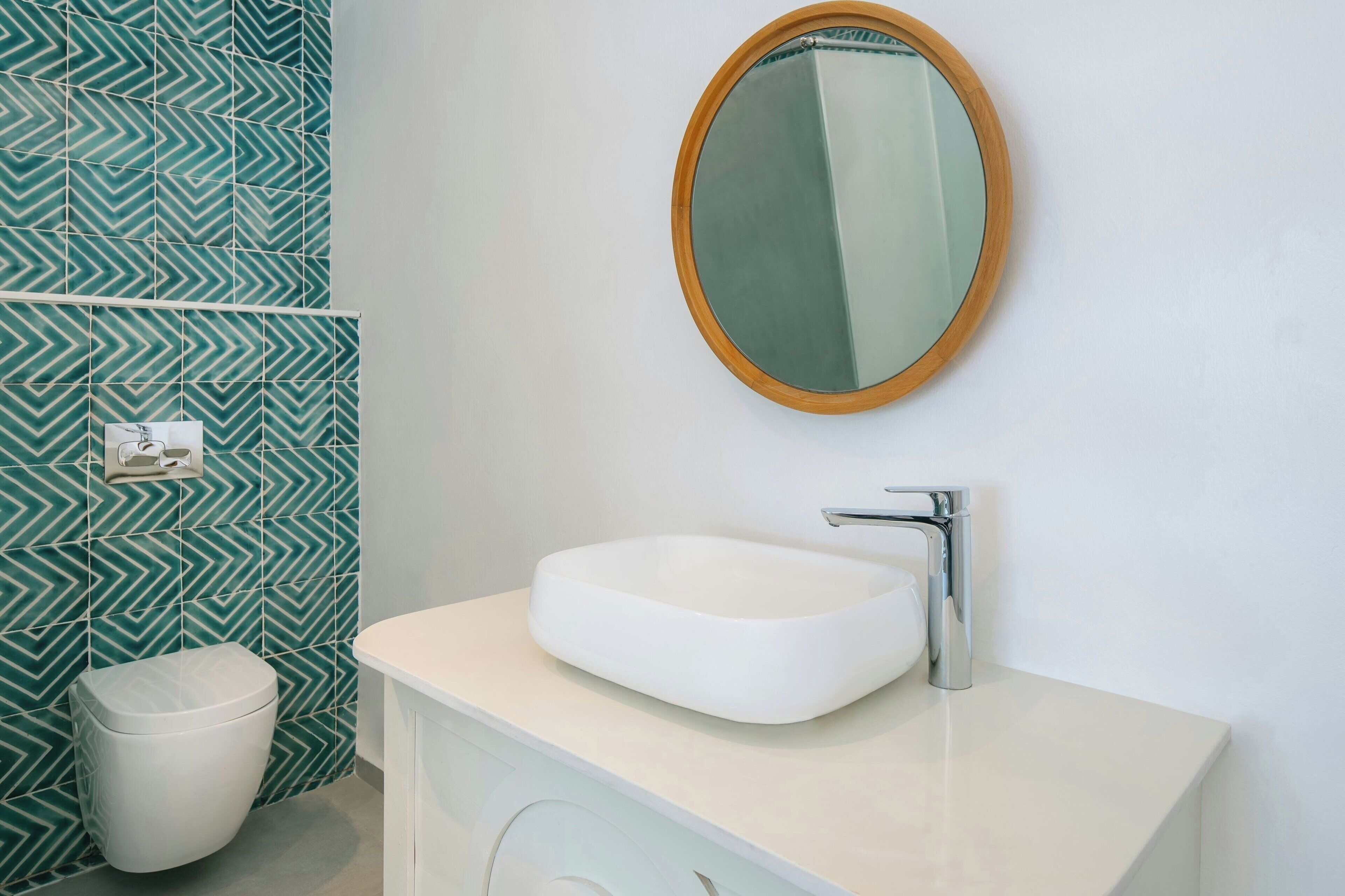 A bathroom with a white sink, a mirror with a wooden frame, and a toilet. The wall has a green and white patterned tile design.