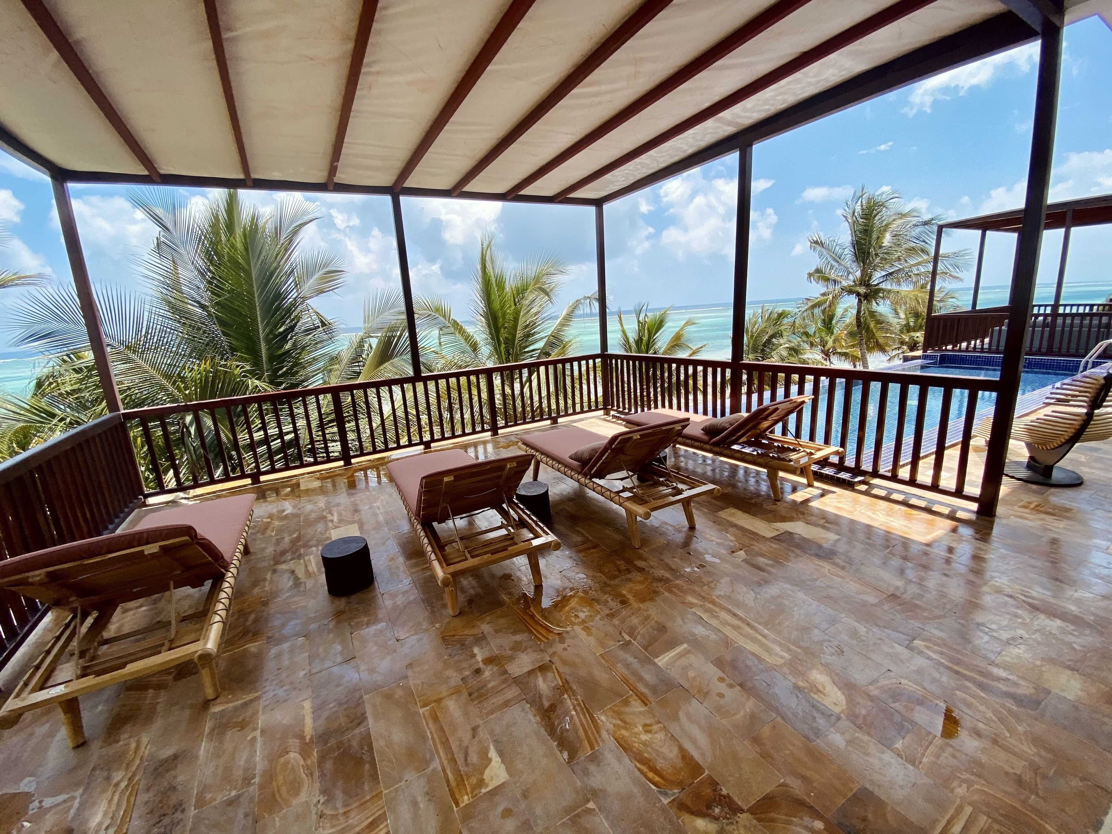 A covered outdoor area with wooden flooring and lounge chairs, overlooking a pool and palm trees.